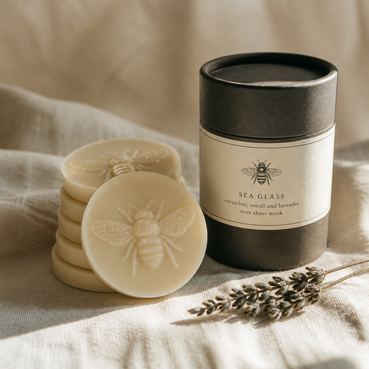Stack of bee-embossed wax medallions beside a charcoal tube on soft linen with dried botanicals